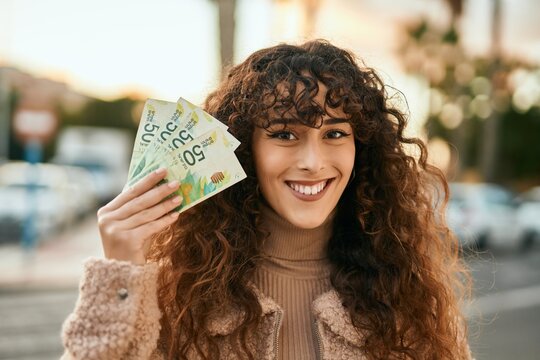 Young hispanic woman smiling happy holding israel shekels banknotes at the city
