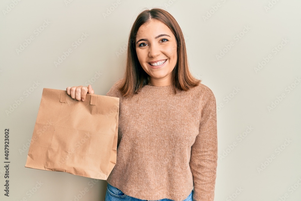 Poster Young beautiful woman holding take away paper bag looking positive and happy standing and smiling with a confident smile showing teeth