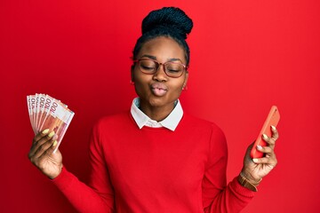 Young african american woman using smartphone holding norwegian krone banknotes looking at the...