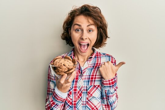 Young brunette woman holding chocolate chips cookies pointing thumb up to the side smiling happy with open mouth