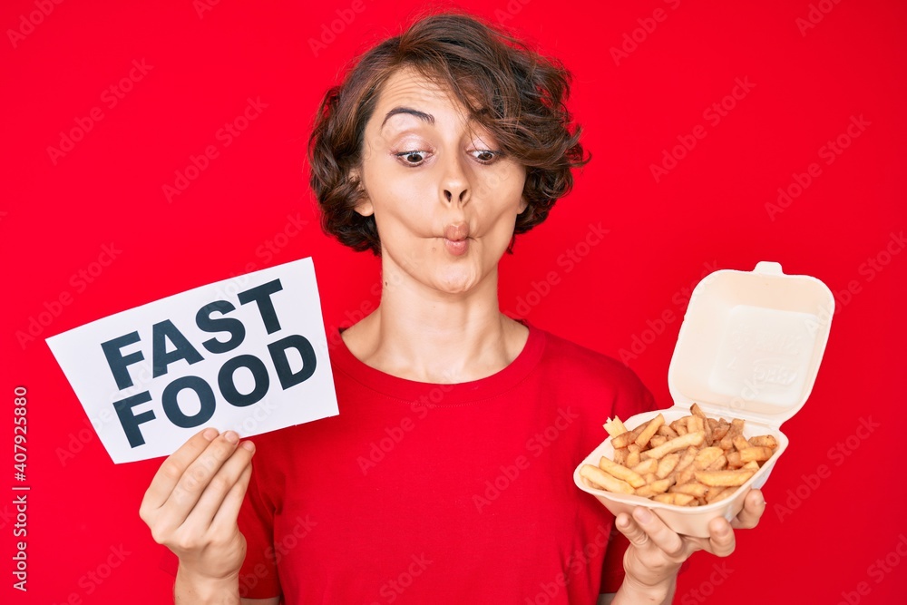 Poster Young hispanic woman holding potato chip and fast food banner making fish face with mouth and squinting eyes, crazy and comical.