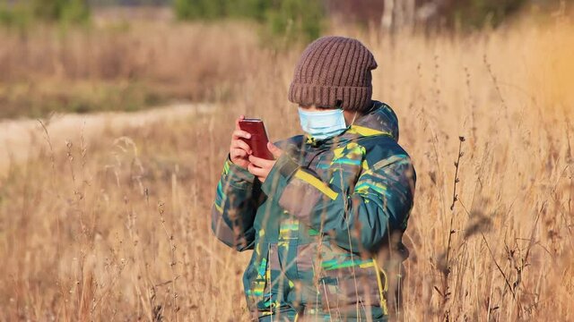 A boy in a medical mask the tall grass, he uses the Internet on his phone. Walk in the park in autumn. Camping on a hike. Healthy lifestyle concept. At sunset, in the evening, warm soft light.