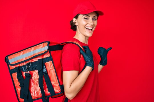 Young hispanic woman holding take away backpack pointing thumb up to the side smiling happy with open mouth