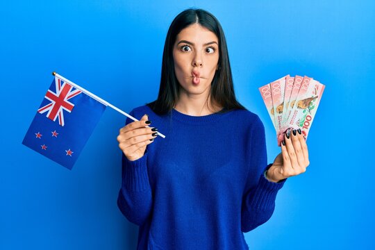 Young hispanic woman holding new zealand flag and dollars making fish face with mouth and squinting eyes, crazy and comical.