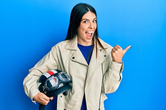 Young hispanic woman holding motorcycle helmet pointing thumb up to the side smiling happy with open mouth
