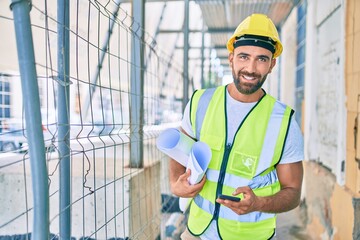 Young hispanic architect man holding blueprint using smartphone at street of city