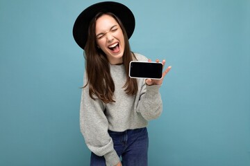 Portrait photo shot of beautiful young woman wearing black hat and grey sweater holding phone showing smartphone isolated on background with open mouth having joy