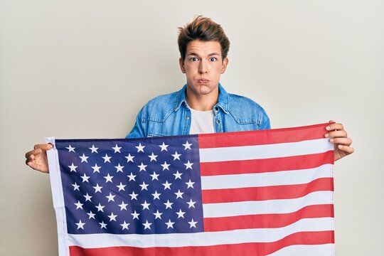 Handsome caucasian man holding united states flag puffing cheeks with funny face. mouth inflated with air, catching air.