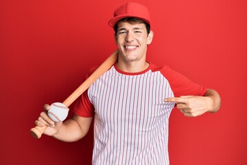 Handsome caucasian man playing baseball holding bat and ball pointing finger to one self smiling...