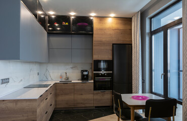 The interior of the studio kitchen is decorated in gray tones with wooden veneers. Kitchen appliances and top cabinets are black. The table top is white. Large panoramic window.