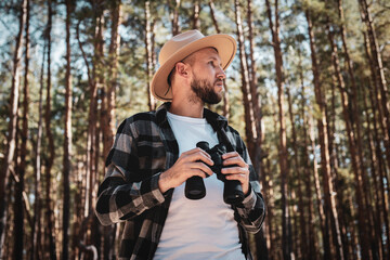 Obraz premium Man tourist in a hat and gray plaid shirt looks through binoculars