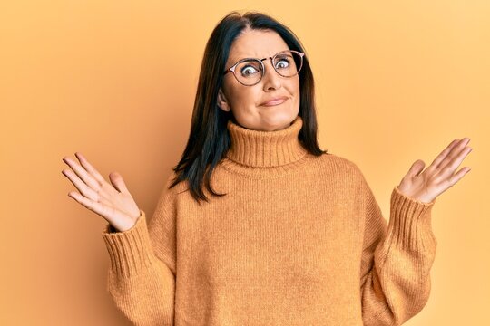 Middle age brunette woman wearing casual winter sweater over yellow background clueless and confused with open arms, no idea and doubtful face.