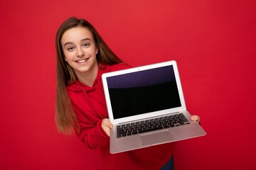 Naklejka premium Close-up portrait photo of beautiful happy smiling girl with long hair wearing red hoodie holding computer laptop looking at camera isolated over red wall background