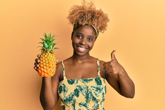 Young african woman with afro hair holding pineapple smiling happy and positive, thumb up doing excellent and approval sign
