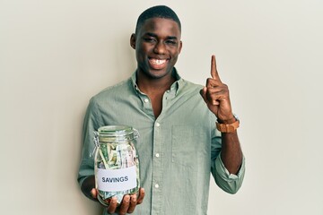 Young african american man holding jar with savings smiling with an idea or question pointing...