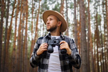 Young Man in a hat with a backpack and binoculars in a pine forest. Hike in the mountains or forest