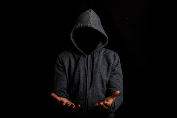 Man without a face in a hood holds something in his hands on a dark background - obrazy, fototapety, plakaty