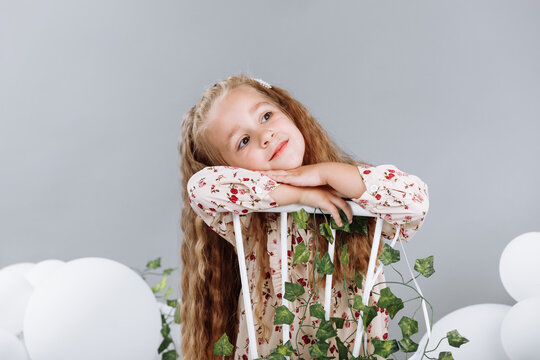 Cute little girl blonde smiling having fun in studio with white balloons and green. child in floral dress sitting on white chair and celebration easter. happy childhood