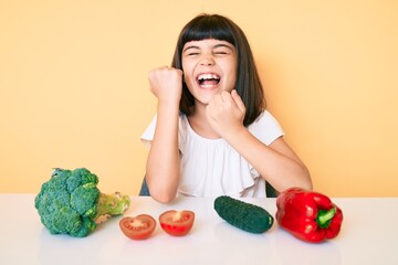 Young little girl with bang sitting on the table with veggies celebrating surprised and amazed for success with arms raised and eyes closed