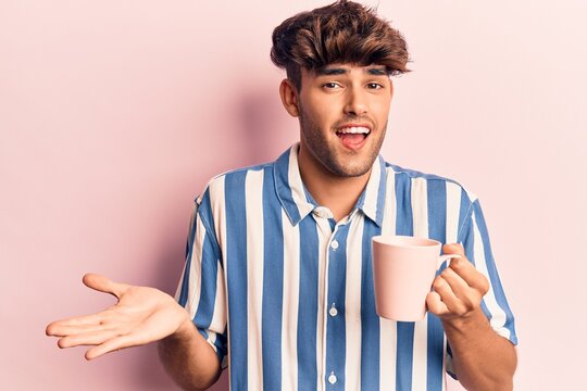 Young hispanic man holding coffee celebrating achievement with happy smile and winner expression with raised hand