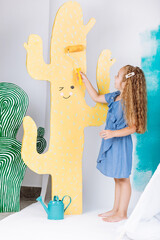 Portrait of a beautiful smiling girl with a paint roller and big paper cactus in studio. Cheerful cute baby having fun in the nursery. baby's day