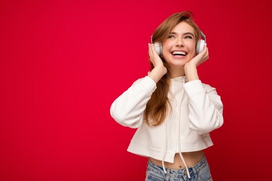 Photo of beautiful happy smiling young blonde woman wearing white hoodie isolated over colourful background wall wearing white wireless bluetooth earphones listening to cool music and enjoying looking