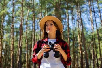 Woman in hat and red plaid shirt holding binoculars on forest background