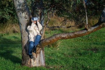 Vista de frente de una joven hispana de cabello negro y rizado que lleva un casco de realidad virtual y disfruta de la experiencia subida en un árbol en el bosque vestida de manera casual en invierno.