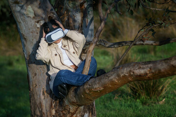 Naklejka premium Vista de frente de una joven hispana de cabello negro y rizado que lleva un casco de realidad virtual y disfruta de la experiencia subida en un árbol en el bosque vestida de manera casual en invierno.