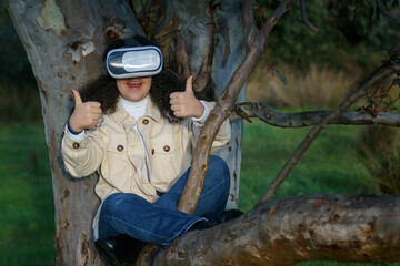 Obraz premium Vista de frente de una joven hispana de cabello negro y rizado que lleva un casco de realidad virtual y disfruta de la experiencia subida en un árbol en el bosque vestida de manera casual en invierno.