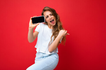Naklejka premium Happy satisfied beautiful smiling young blonde woman good looking wearing white t-shirt standing isolated on red background with copy space holding phone showing smartphone in hand with empty screen