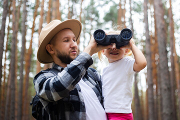 Man in a hat and a backpack and a child look through binoculars while hiking in the forest. Family hike to the mountains or forest