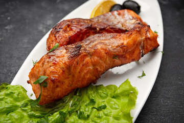 Two baked grilled pieces of salmon fish on a white plate with lettuce, olives and lemon on a dark background