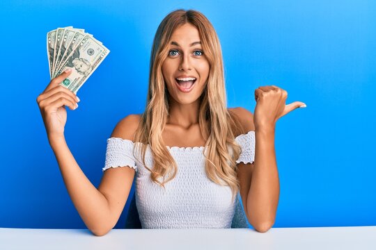 Beautiful blonde young woman holding 20 dollars banknotes pointing thumb up to the side smiling happy with open mouth