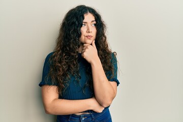 Young brunette woman with curly hair wearing casual clothes with hand on chin thinking about question, pensive expression. smiling with thoughtful face. doubt concept.