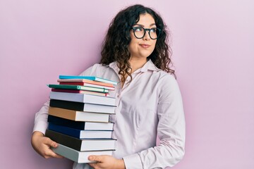 Young brunette woman with curly hair holding a pile of books smiling looking to the side and staring away thinking.