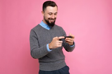 Shot of happy handsome young brunette unshaven man with beard wearing everyday grey sweater and blue shirt isolated on background wall holding smartphone playing games via mobile phone looking at