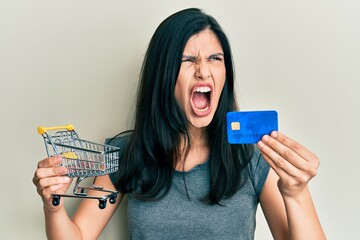 Young hispanic woman holding small supermarket shopping cart and credit card angry and mad screaming frustrated and furious, shouting with anger. rage and aggressive concept.