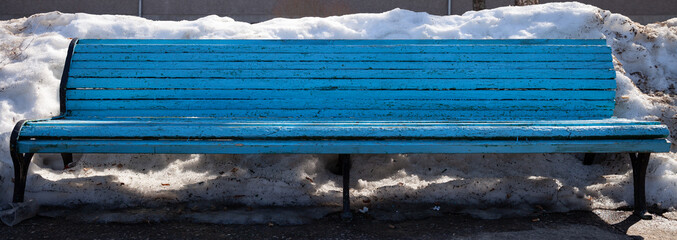 A bright blue, old wooden bench outside in the city on a warm spring day. Painted wood. The beginning of spring. Melting snow drifts. Some rubbish under the bench. Wide view