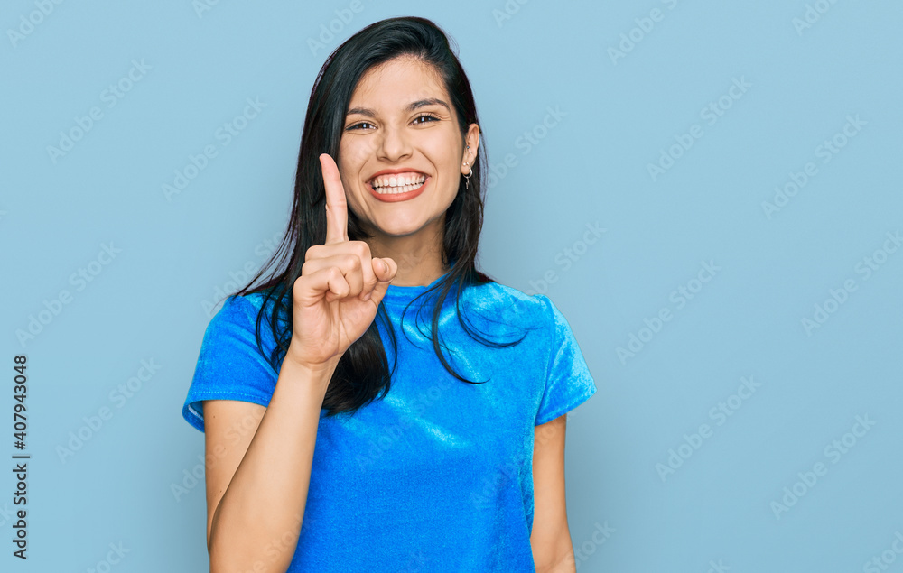 Wall mural young hispanic woman wearing casual clothes showing and pointing up with finger number one while smi