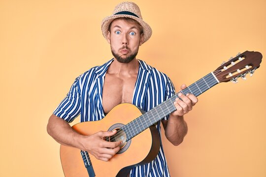 Young caucasian man wearing summer style playing classical guitar puffing cheeks with funny face. mouth inflated with air, catching air.