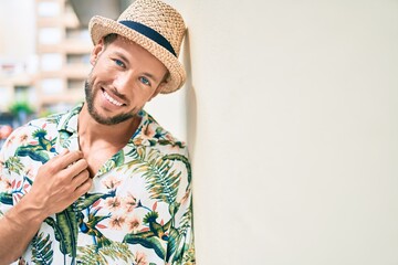 Handsome caucasian man wearing summer hat and flowers shirt smiling happy leaning on the wall