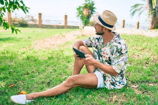 Caucasian handsome man smiling happy using touchpad device sitting on the grass at the park