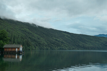 A wooden cottage located at the shore of Millstaetter lake in Austria. The lake is surrounded by high Alps. Calm surface of the lake reflecting the mountains. A bit of overcast. Calmness and serenity