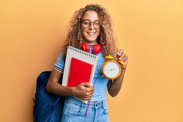 Beautiful caucasian teenager girl wearing student backpack and holding alarm clock winking looking at the camera with sexy expression, cheerful and happy face.