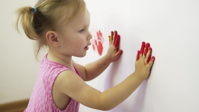 Little beautiful girl paints her handprints with red paint on a white wall.Slow motion.Happy child paints with skin-safe paints.