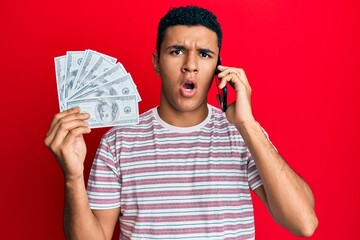 Young arab man having conversation talking on the smartphone holding dollars in shock face, looking...