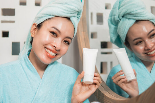 Beautiful Asian Woman Holding Tube Of Facial Foam, Cream Or Lotion In Front Of Mirror Bathroom. Skincare And Beauty Concept.