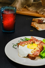 Open sandwich with bacon, fried egg and celery leaves on a slice of rye sourdough bread on a white plate. Glass with fruit drink on the table. Healthy food concept, traditional craft bread. Close-up