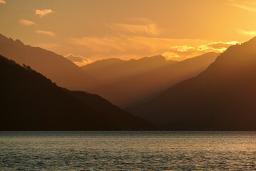 A sunset by Millstaetter lake in Austria. The lake is surrounded by high Alps. Calm surface of the lake reflecting the sunbeams. The sun sets behind the mountains. A bit of overcast. Natural beauty